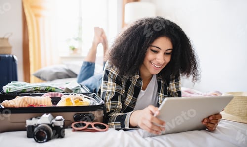 Preview: Young woman with suitcase packing for holiday at home, using tablet