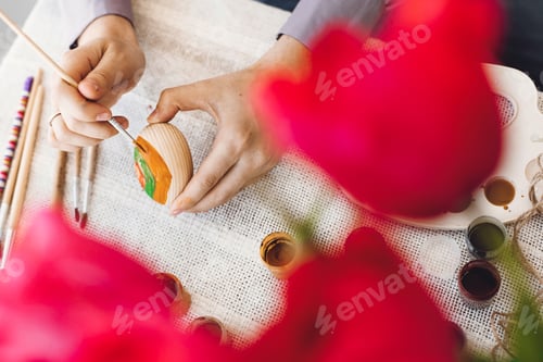 Preview: Hands holding and painting easter egg on rustic table with paint, brushes, tulips bouquet