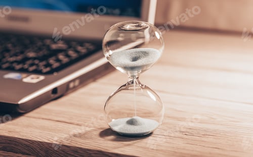 Preview: Hourglass on a light wooden table with a laptop or computer included. Time slips through my fingers.