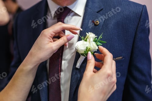 Preview: bride puts groom on boutonniere from pink and whote rose on wedding day