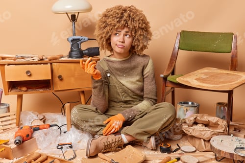 Preview: Consfused curly woman prepares materials for producing wooden furniture processes wood wears