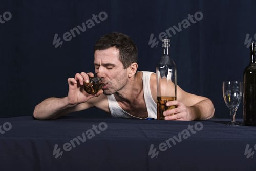 Preview: Drunken lonely man lies with his chest on table and drinks alcohol from glass.