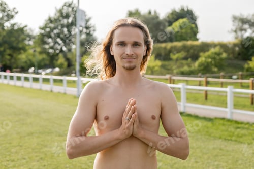 Preview: long haired and shirtless man smiling at camera while meditating with anjali mudra gesture outdoors