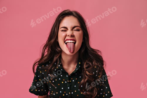 Preview: Portrait of joyful girl showing her tongue on pink background. Happy girl in black polka-dot clothe