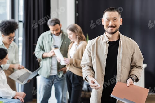 Preview: cheerful asian man holding smartphone and folder near interracial team on blurred background
