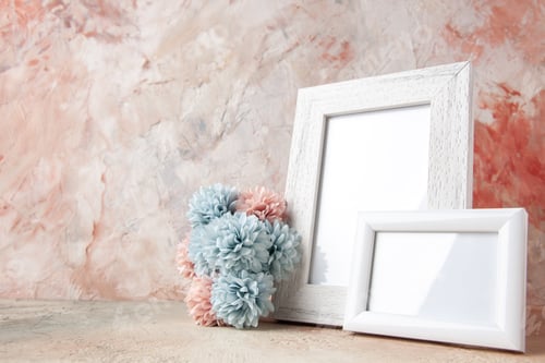 Preview: Close up view of white empty wooden photo frame and flower on the left side on pastel colors