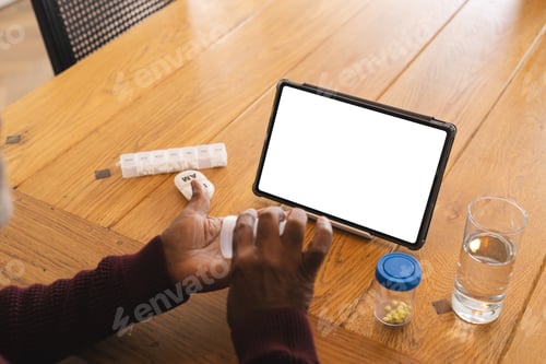 Preview: Senior biracial man having video call with doctor on tablet with copy space, holding pills at home