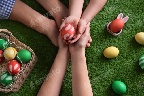 Preview: Father, mother and their child holding painted Easter egg on green grass, top view