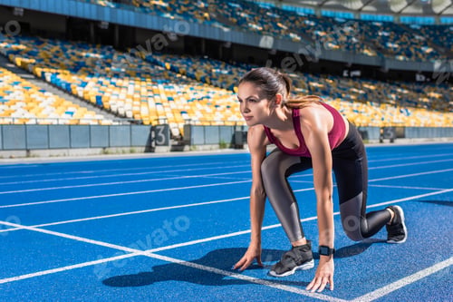 Preview: fit female runner in start position on running track at sports stadium