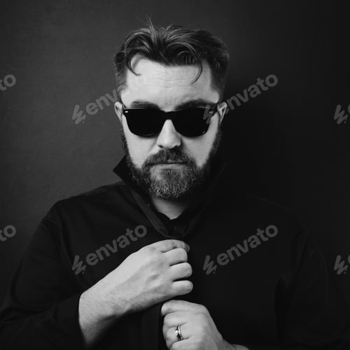 Preview: A brutal man with a beard and a stylish hairstyle in a black shirt adjusts his tie.
