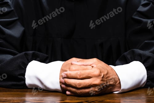 Preview: cropped view of judge in judicial robe sitting at table with clenched hands isolated on black