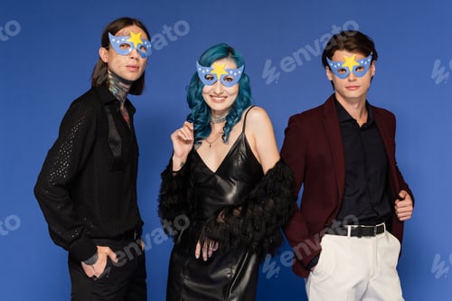 Preview: nonbinary friends in party masks standing with hands in pockets near queer person in black leather