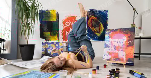 Preview: A woman artist lies on her back on a white tiled floor, surrounded by art supplies and canvases. Pai
