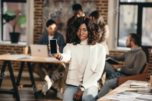 Preview: serious african american casual businesswoman pointing at smartphone with blank screen in loft