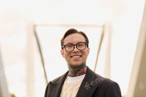 Preview: Handsome young man with tattoos showcases stylish attire against modern architectural backdrop