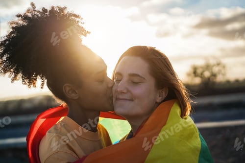Preview: Happy women gay couple kissing and wearing rainbow flag outdoor - Love and lesbian concept