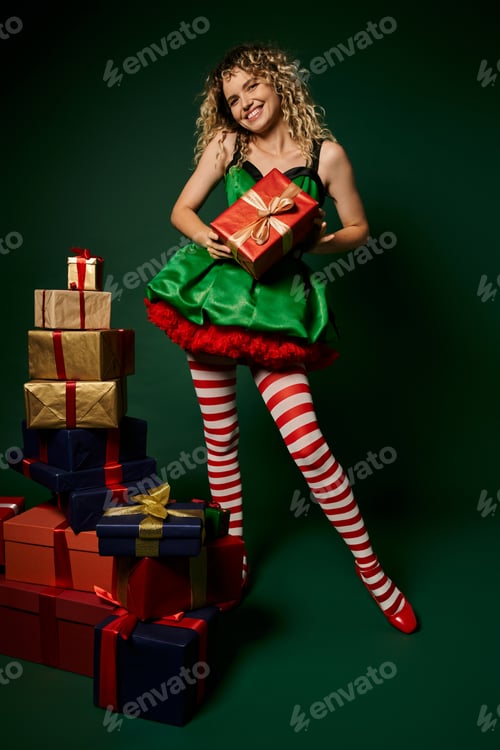 Preview: pretty curly woman in green dress posing with presents on green backdrop, new year elf concept