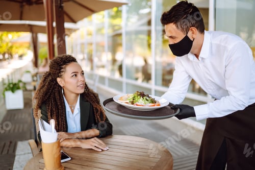 Preview: Young waiter in protective face mask and gloves while bringing food to a customer in cafe. Covid-19.