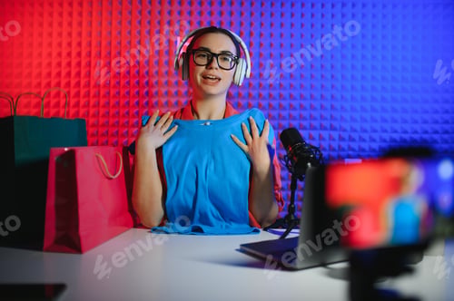 Preview: Positive trendy blogger woman speaking at smartphone webcam on tripod, recording post for vlog