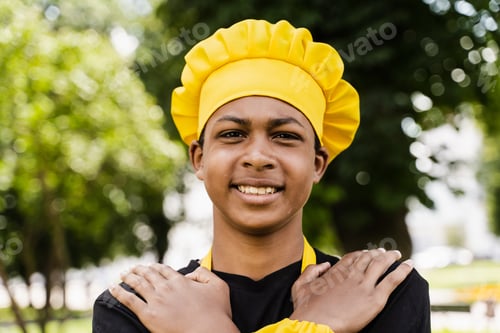 Preview: Black teen chef smiling outdoors, yellow uniform, restaurant promotional photo