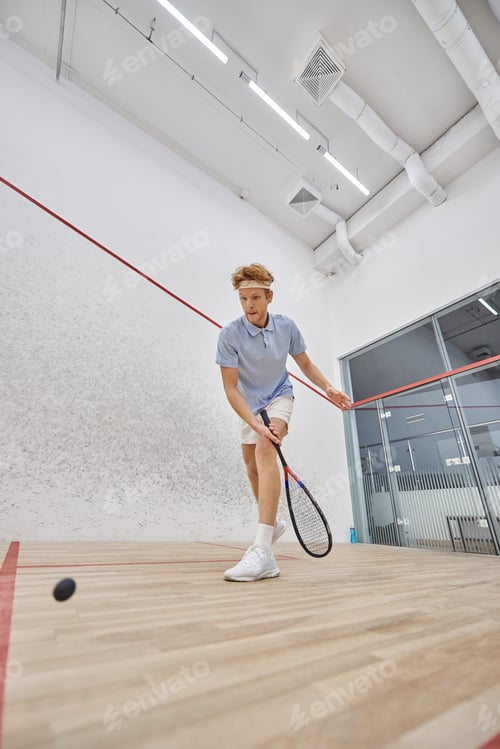 Preview: active redhead sportsman in headband playing squash inside of court, challenge and motivation