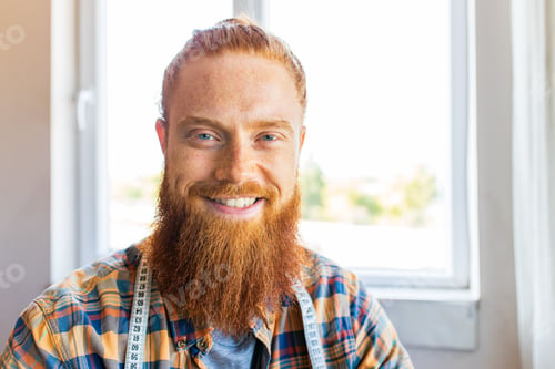 Preview: handsome redhaired man with long beard sews at a sewing machine at home studio