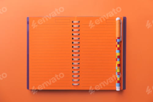 Preview: A Vibrant Orange Notebook is Open with a Colorful Pen Laying on a Light Background Surface
