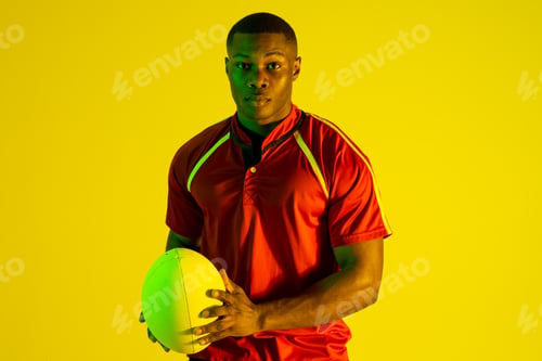 Preview: Portrait of african american male rugby player with rugby ball over yellow lighting