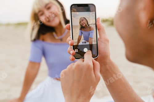 Preview: Young man taking photo of his girlfriend during picnic on beach