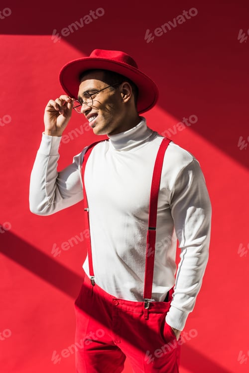 Preview: fashionable mixed race man in hat and suspenders smiling and posing on red