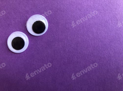 Preview: Googly eyes on a plain purple textured background with copy space. Creative fun photography