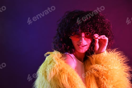Preview: Beautiful young woman in yellow fur coat and sunglasses on color background in neon lights