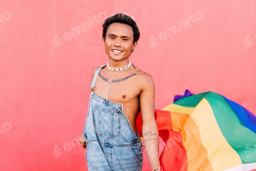 Preview: Smiling guy holding a rainbow LGBT flag while standing against a pink wall outdoors