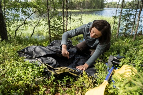 Preview: Woman unfolds new rubber boat, oars lying nearby, against of lake