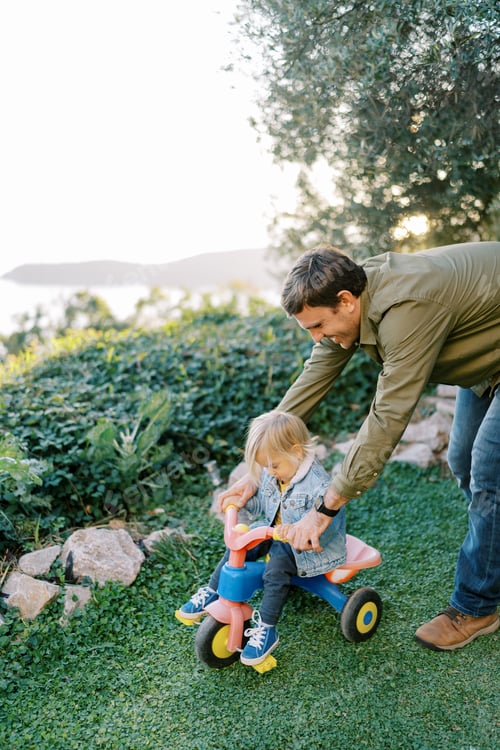 Preview: Smiling dad pushes a little girl riding a tricycle in the park
