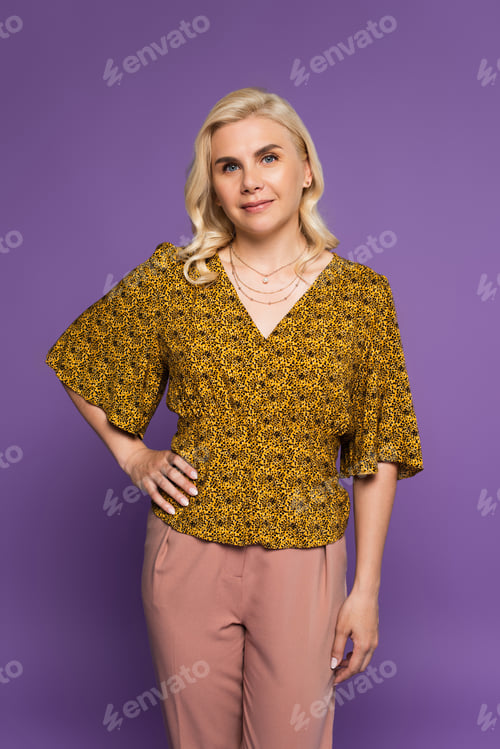 Preview: happy and blonde woman in blouse posing with hand on hip isolated on purple