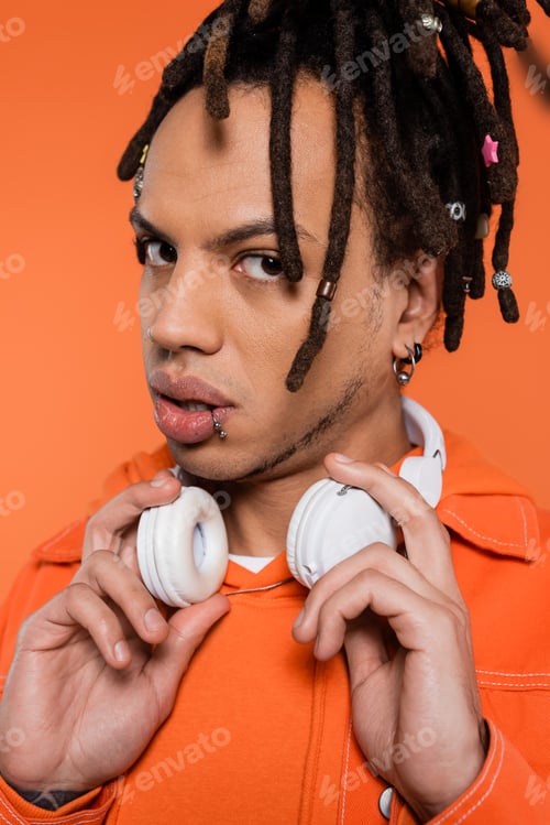 Preview: tattooed and multiracial man with wireless headphones looking at camera isolated on coral background
