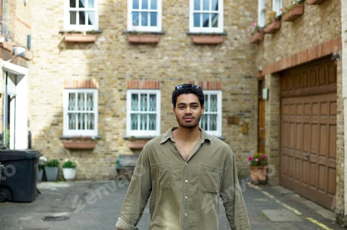 Preview: Young man standing in front of a brick house lined with windows and adorned with plants.