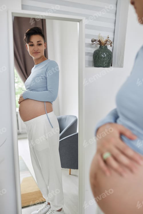 Preview: Young pregnant woman looking in mirror measuring baby bump
