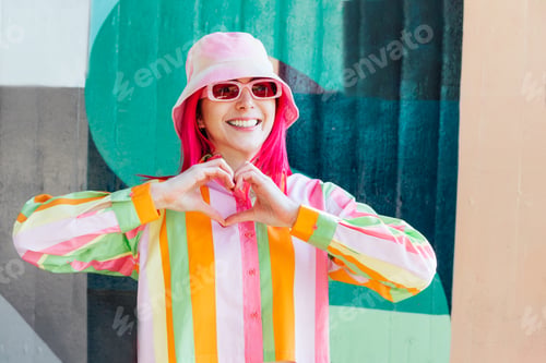 Preview: Young woman with pink hair and sun glasses in bucket hat and multicolor shirt making sign Heart