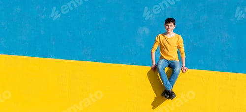 Preview: Young boy wearing casual clothes sitting on a yellow fence against a blue wall in a sunny day