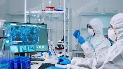 Preview: Doctor in ppe suit working at pc while chemist using microscope