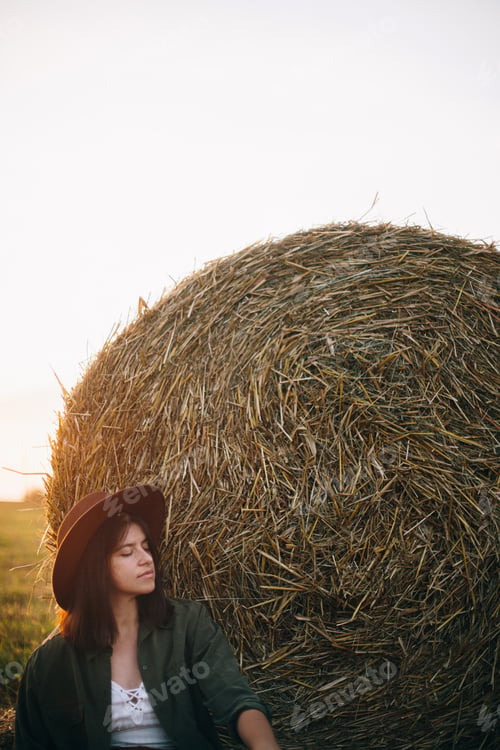 Preview: Beautiful woman in hat relaxing at haystacks enjoying evening sunset in summer field. Tranquility