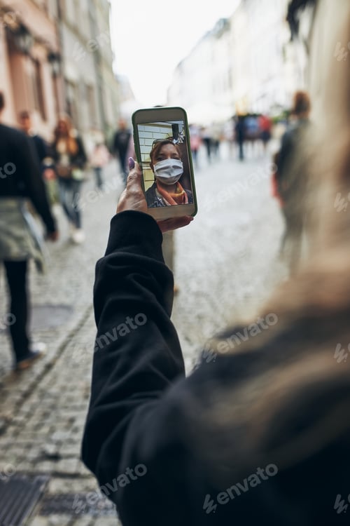 Preview: Young woman having video call talking while walking downtown in the evening wearing the face mask