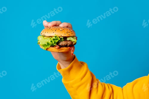 Preview: Male hand in yellow clothes hold hamburger on blue background