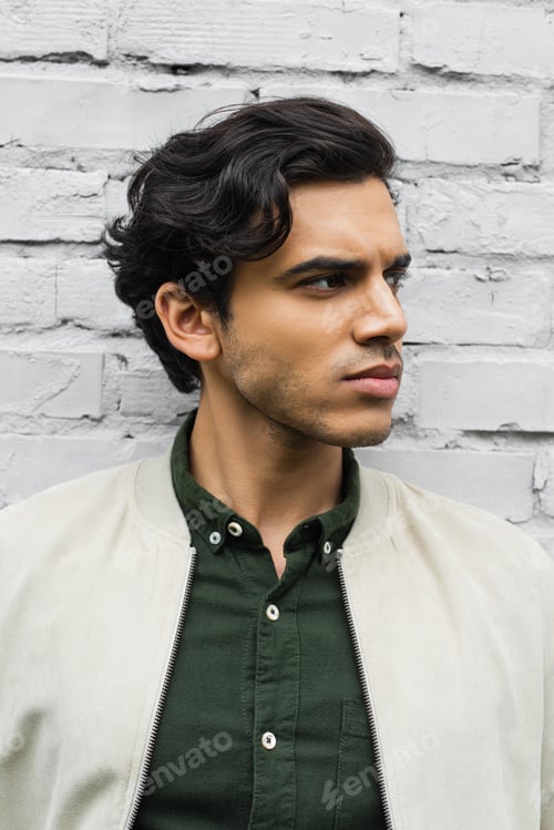 Preview: young man in bomber jacket looking away near brick wall