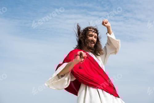 Preview: low angle view of Jesus in robe, red sash and crown of thorns having fun and jumping with rosary