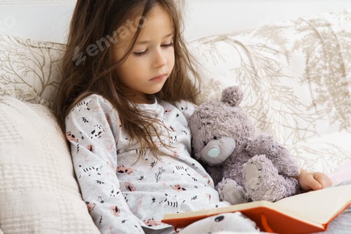 Preview: Little cute, adorable, nice brunette girl in pajamas reading book with teddy bear on soft bed. Learn
