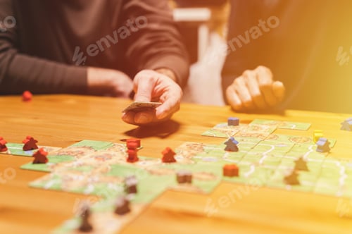 Preview: men friends have fun playing the carcassonne board game at night. male hands and game cards and chip