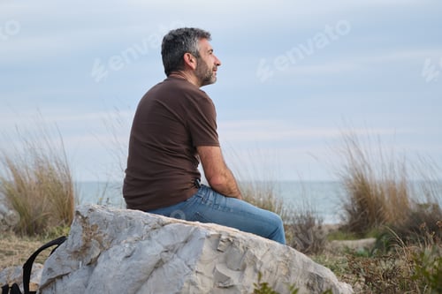 Preview: medium shot of Mature man sitting rock by ocean thinking